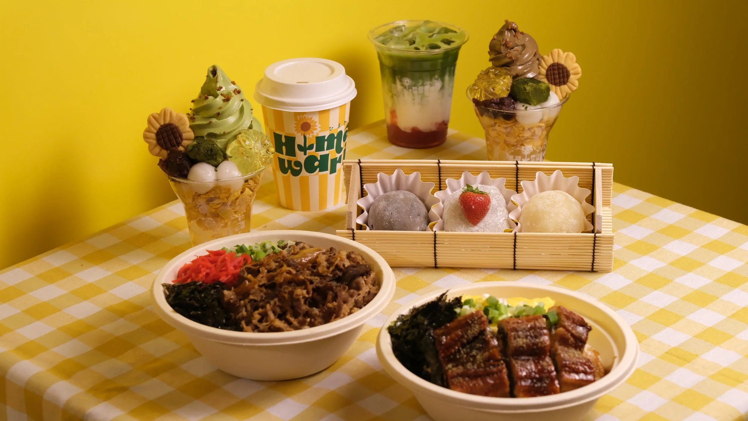 A table with Japanese food and desserts featuring bowls of rice with beef and eel, green tea soft serve, mochi sweets, and parfaits with fruit and toppings, with a yellow and white checkered tablecloth.
