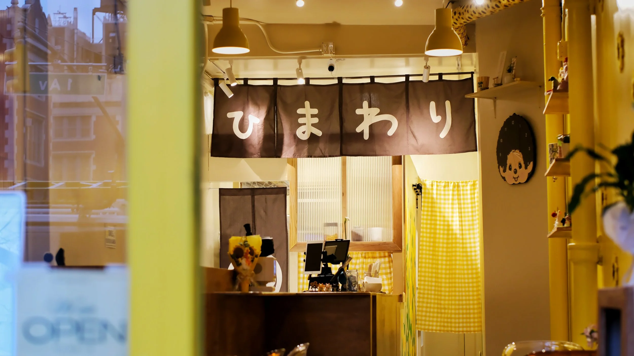 Interior of a cozy shop or cafe with yellow accents, yellow checkered curtains, and decorative wall art including a mermaid image. A brown curtain with Japanese characters hangs above the counter.