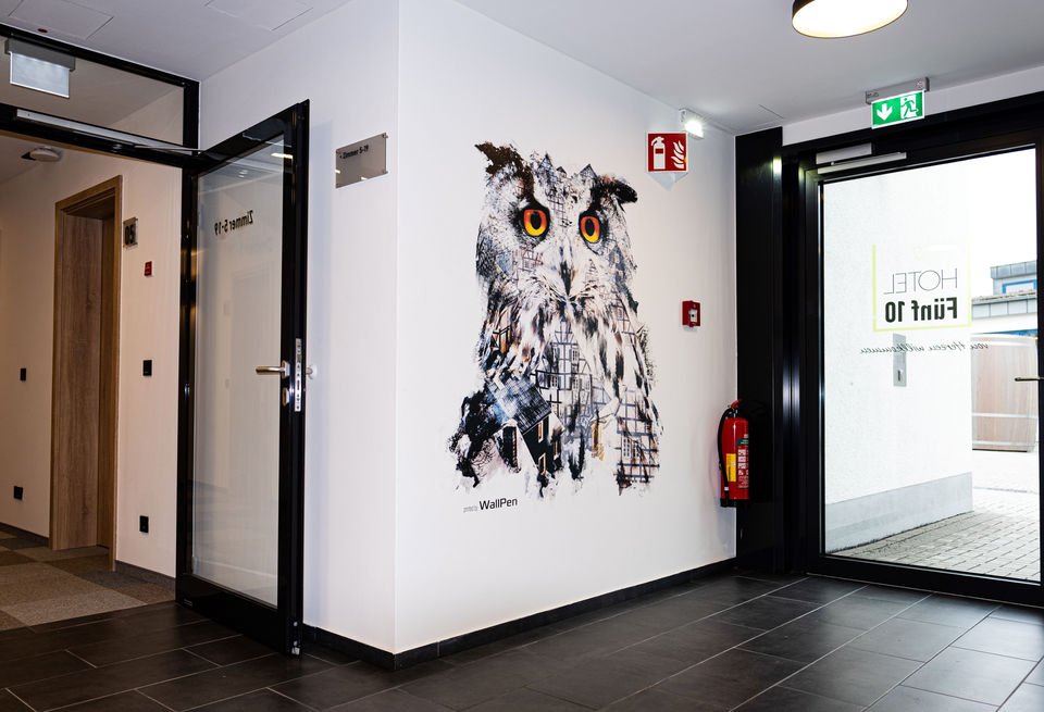 Interior of a hotel hallway with a large owl mural on the wall, an open glass door, a fire extinguisher, and an emergency exit sign.