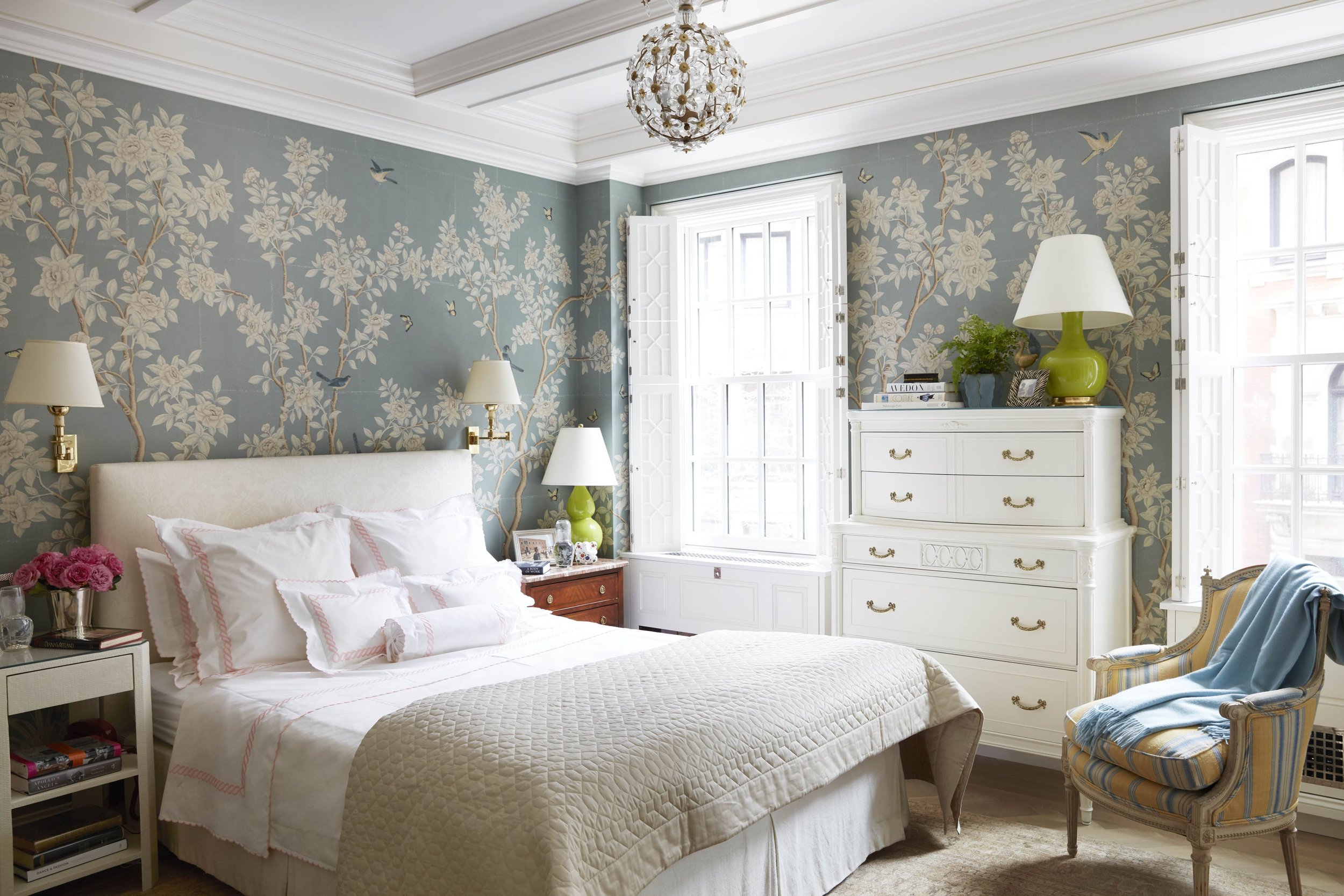 Elegant bedroom with floral wallpaper, white bed with pink-trimmed pillows, white dresser, and vintage chair with blue and yellow striped fabric.