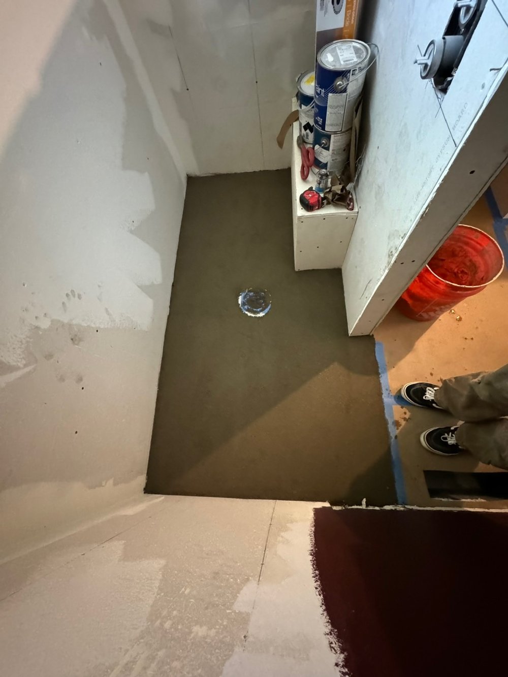 Interior of a room under construction with freshly poured concrete floor, a small hole in the floor, construction tools, and a partially painted wall