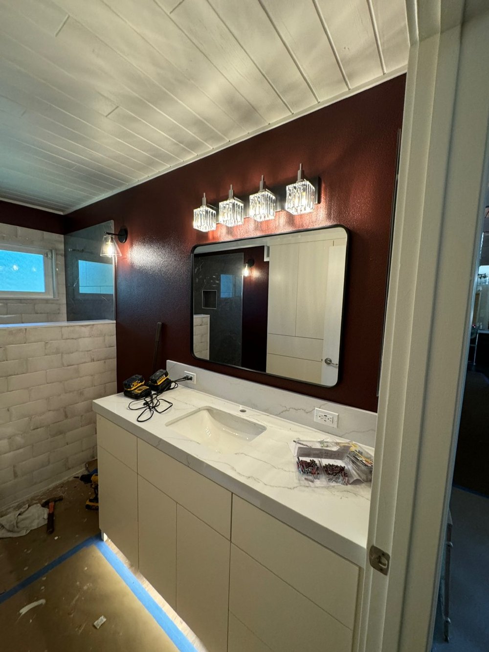 Bathroom vanity with marble countertop, large mirror, and modern light fixture on a dark red wall, under construction with tools and materials nearby.