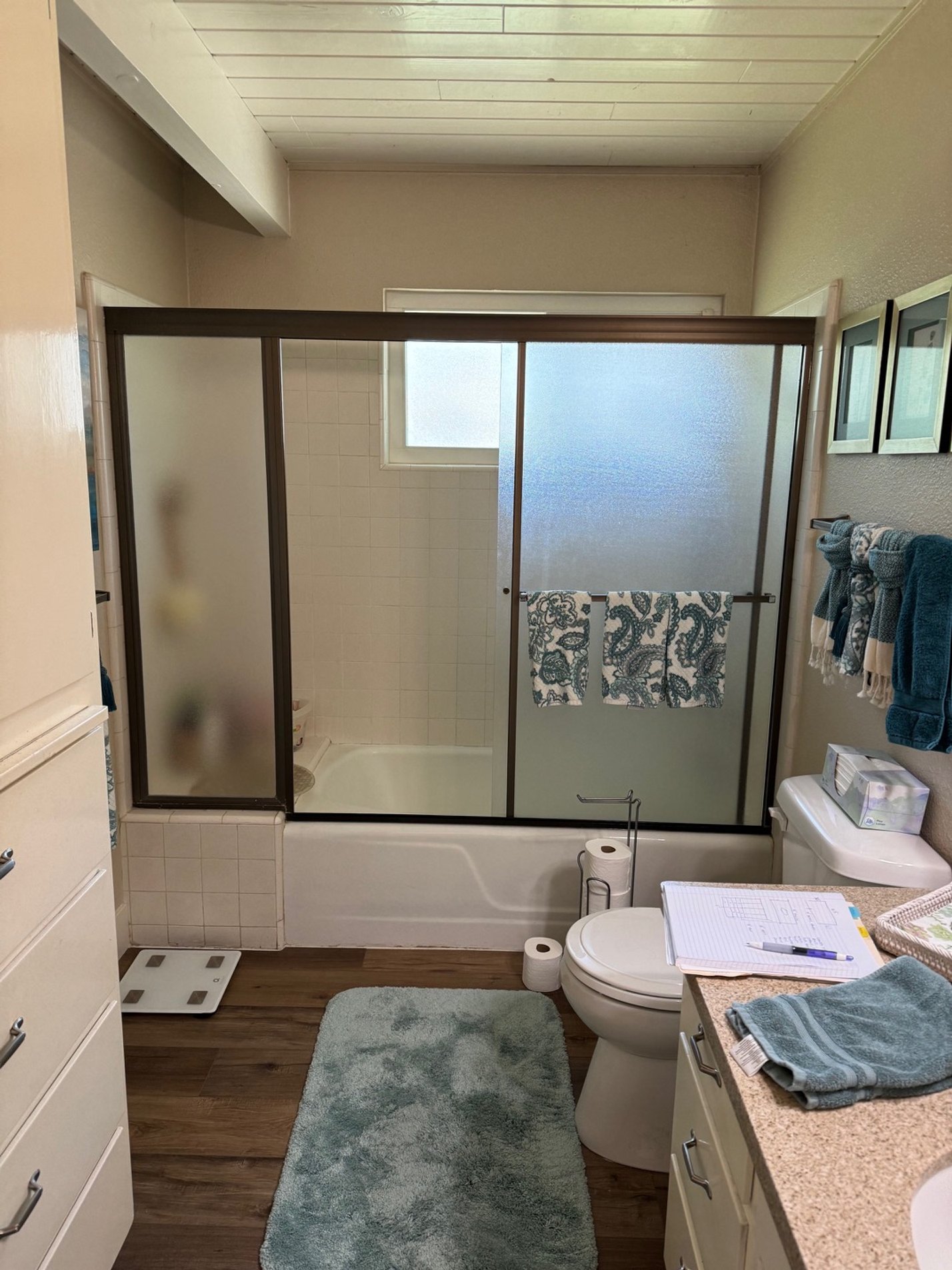 A small bathroom with a bathtub and frosted glass sliding shower door, a toilet, a beige countertop with a blue towel, notebooks, and a pen, and a towel rack with blue and patterned towels; a bathroom scale is on the floor and a bath mat by the toile