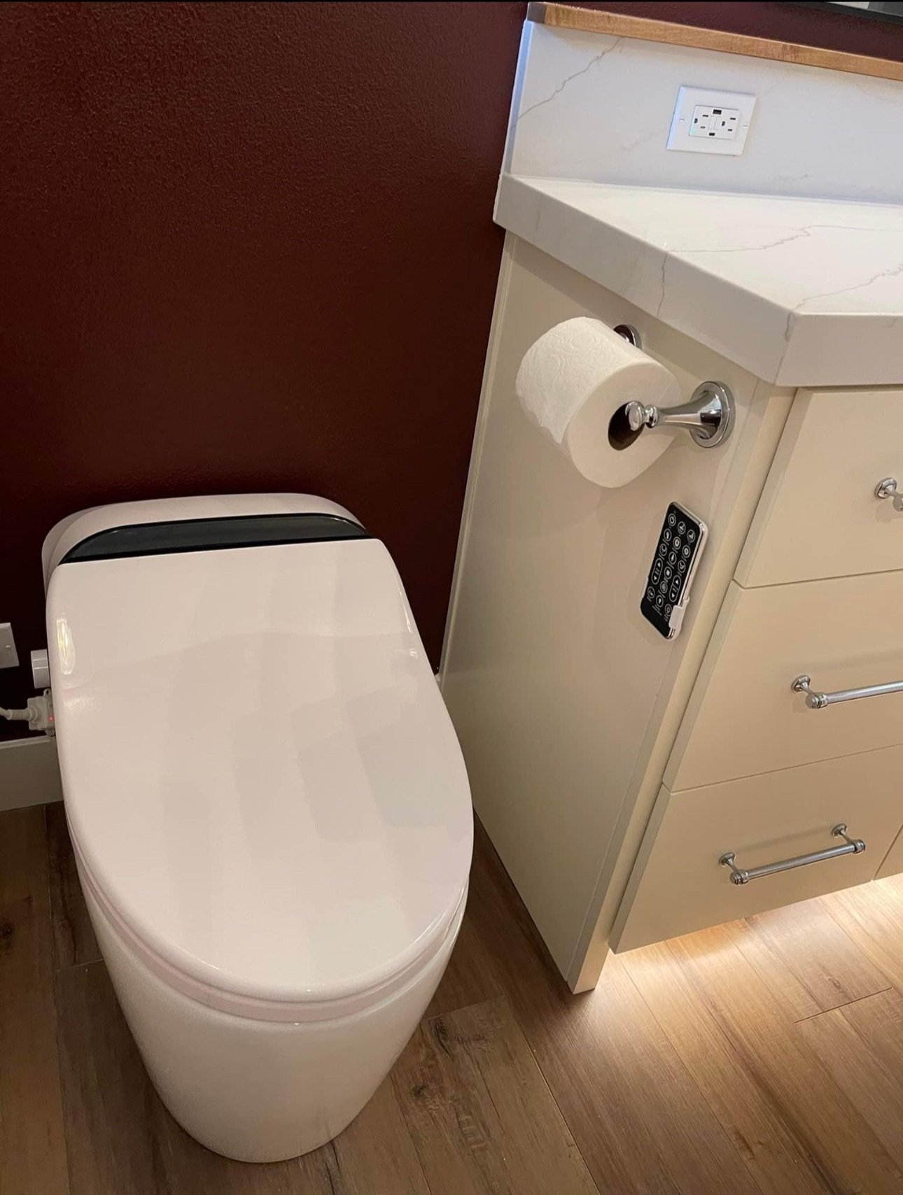A modern American toilet next to a beige cabinet with a roll of toilet paper on a silver holder, a TV remote, and a marble countertop in a bathroom. The wall behind is dark red.