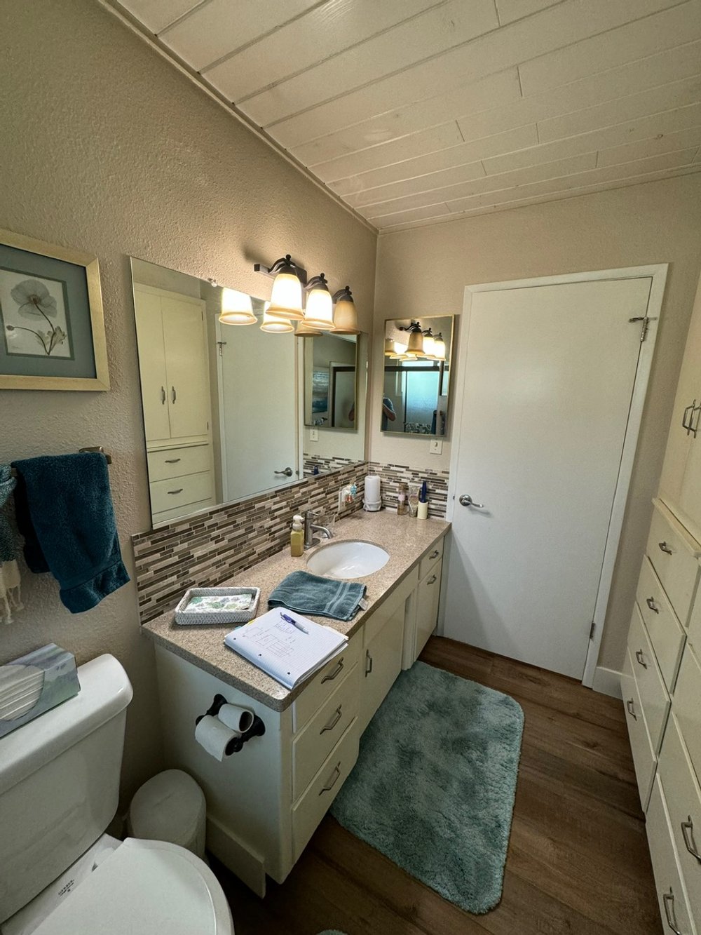A small bathroom with a beige countertop, a sink, a mirror, and a lighting fixture above. There are toilet paper rolls, towels, and toiletries on the counter. The bathroom has a green rug on a wooden floor and white cabinets.