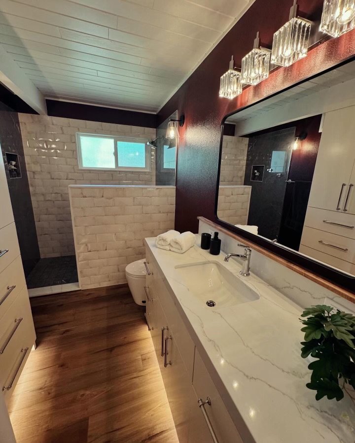 Modern bathroom with a white vanity and marble countertop, large mirror, glass lighting fixtures, beige wood flooring, and a shower area with brick wall dividers and frosted window.