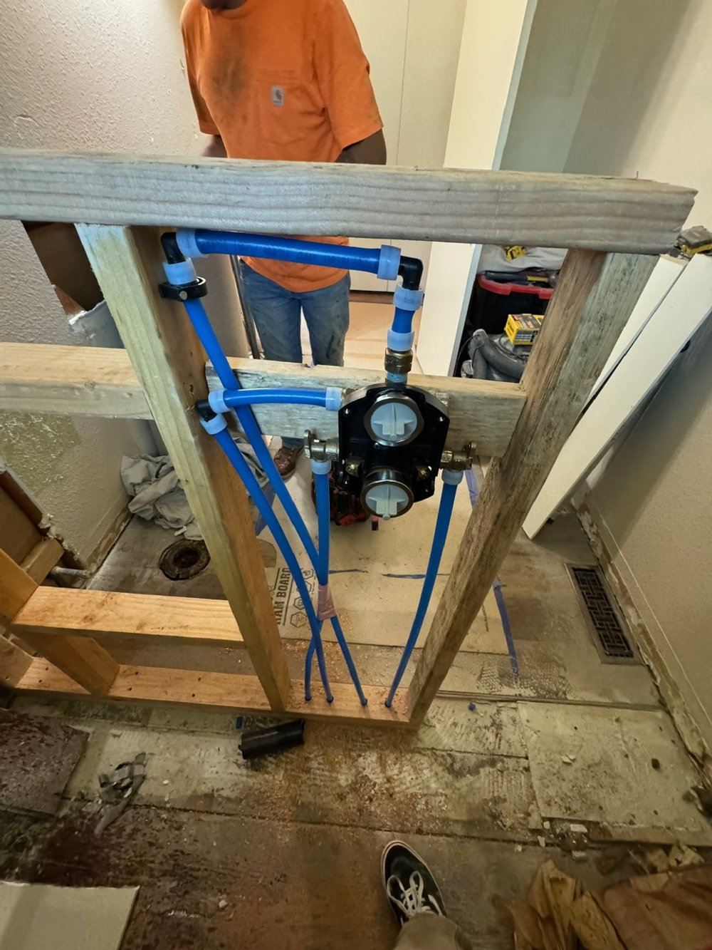 Wall framing with plumbing rough-in in progress, including blue PEX pipes and a black valve or meter, with a person wearing an orange shirt in the background.