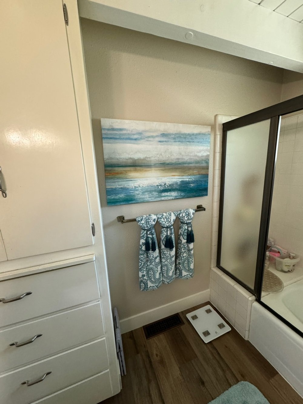 Bathroom corner with white cabinet, abstract ocean painting, patterned towels hanging on a rod, and a digital bathroom scale on wood floor