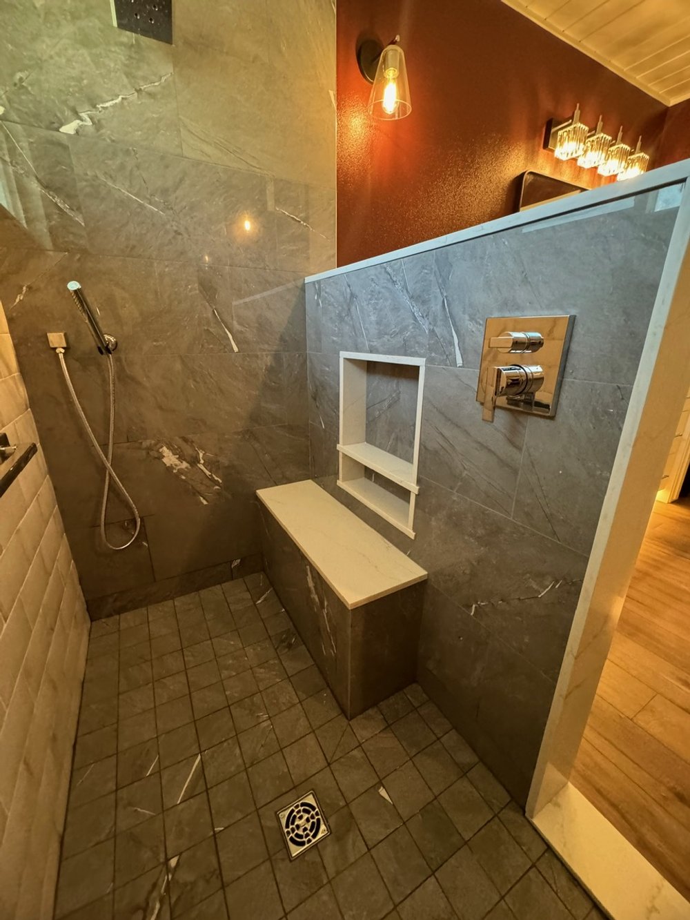 Modern shower area with gray marble walls, a handheld showerhead, built-in shelf, and wooden bench on dark tiled floor.