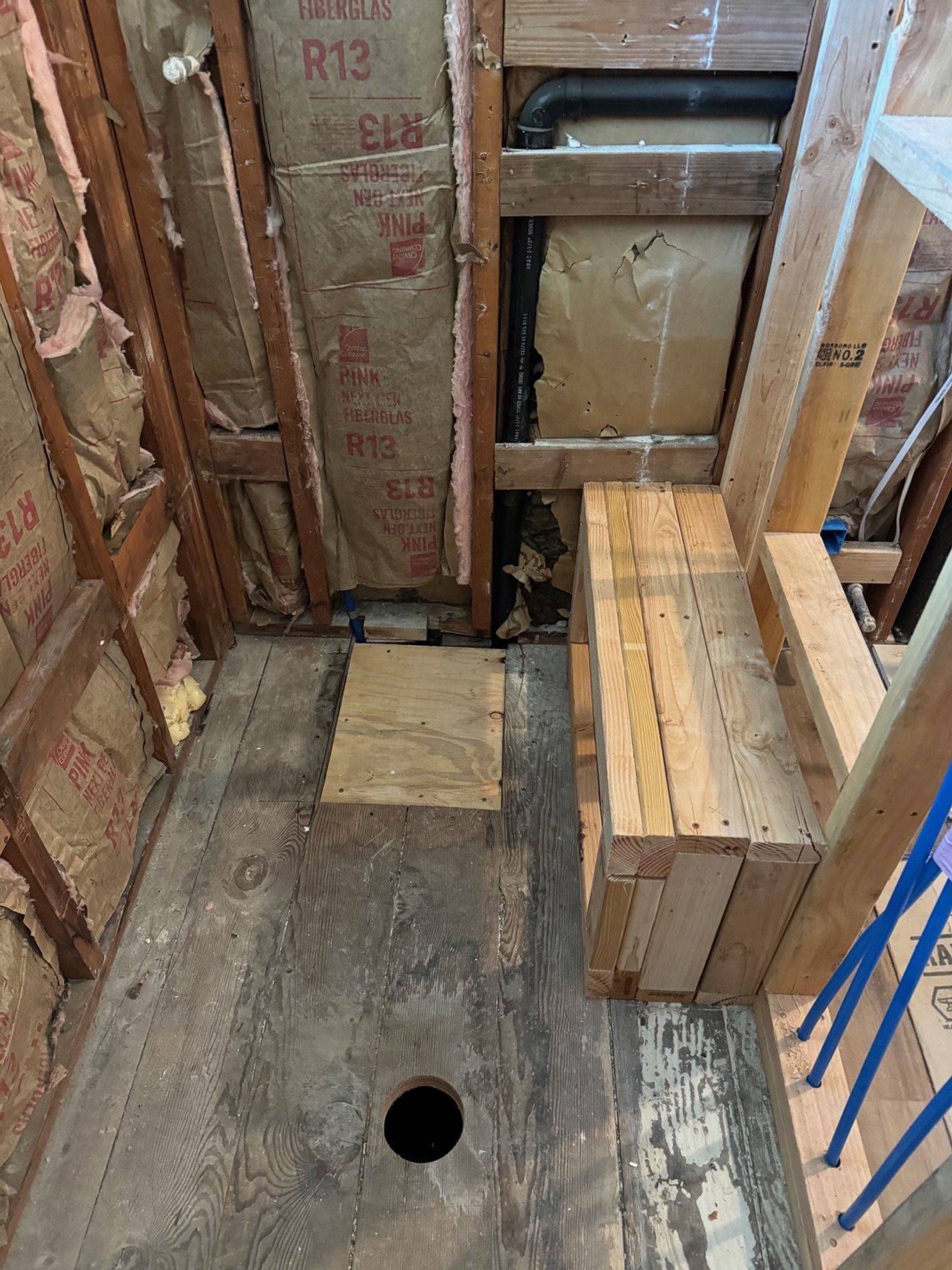 Construction site with exposed wall studs, insulation, plumbing pipes, and unfinished wooden flooring with a hole in it.