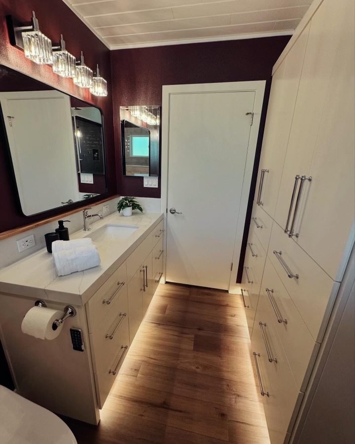 Bathroom with a white vanity, mirror, small plant, rolled white towel, and a soap dispenser, with dark red walls, a door, and large white cabinets, wooden flooring, and modern lighting fixtures.