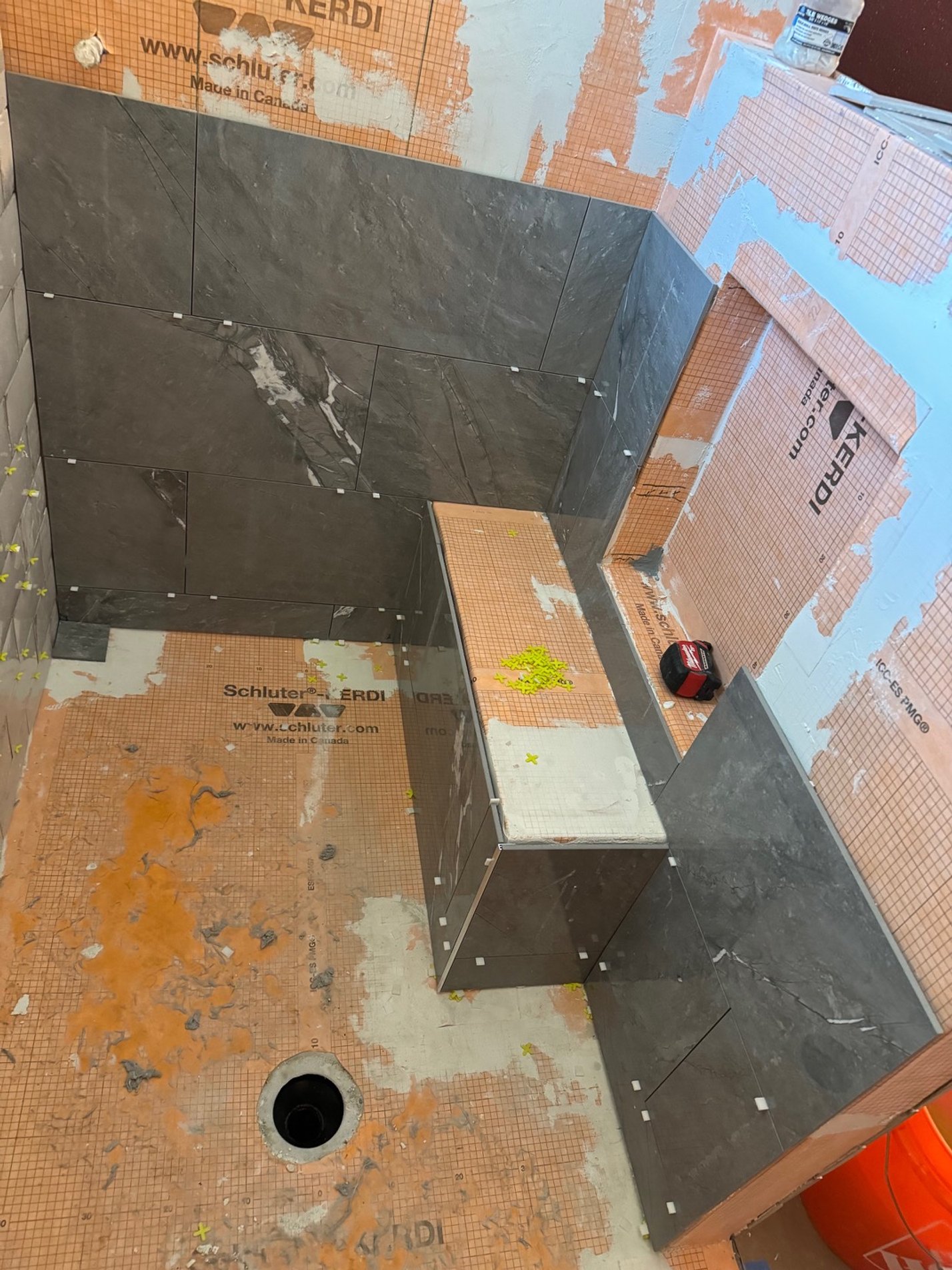Bathroom under construction with dark gray tiles partially installed on walls and orange waterproof membrane on floor and walls, with construction tools and materials present.