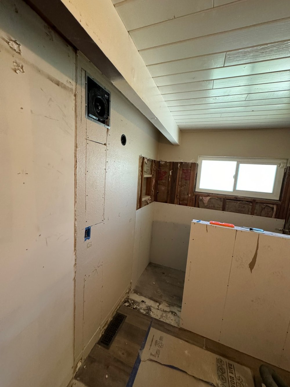 Interior of a room under renovation with unpainted drywall, exposed insulation, and a window. There is a small, partially enclosed area with a drywall panel, a vent on the floor, and construction tools on a ledge.