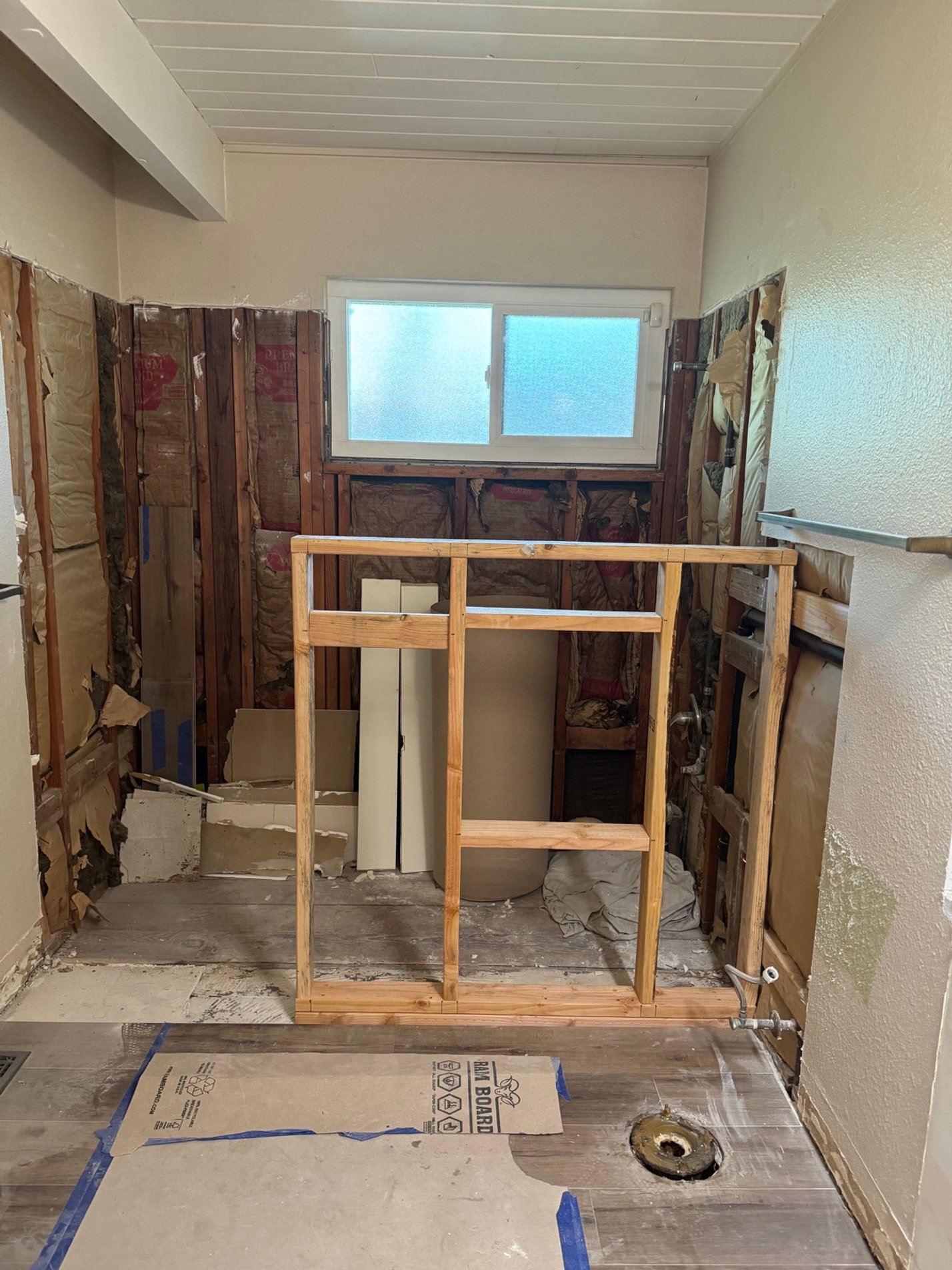 Bathroom under renovation with exposed wall studs, insulation, and a plumbing pipe hole in the floor, a small window at the back, and a wooden frame in the center.