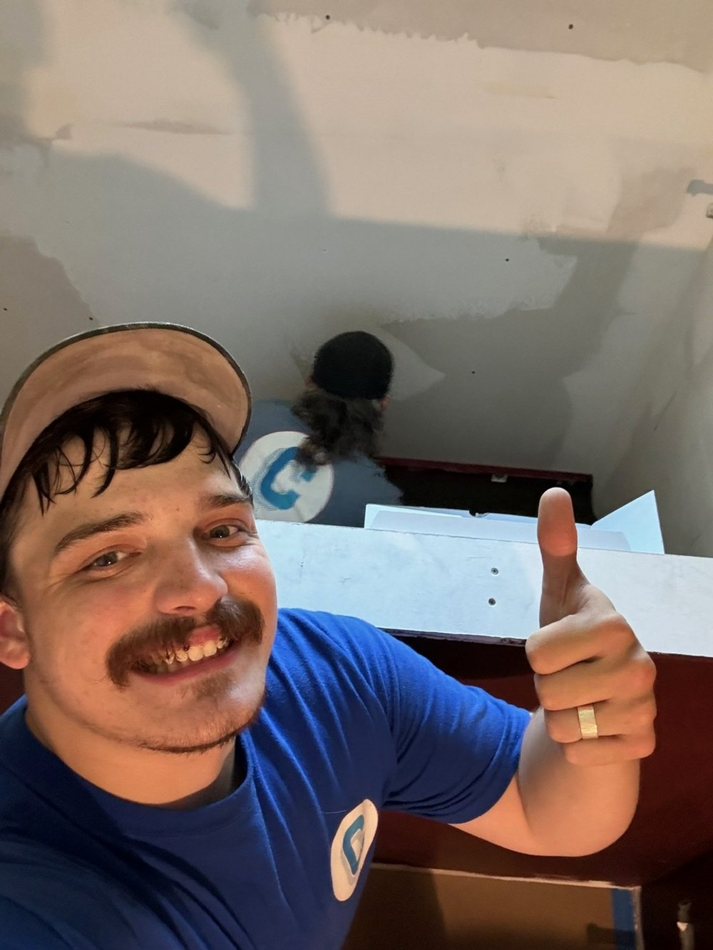 A man smiling and giving a thumbs up, wearing a blue shirt and a tan cap, standing against a stairwell with a person going down the stairs behind him.