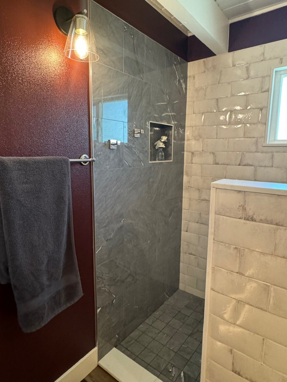 Bathroom shower area with gray tiled wall, a small built-in shelf with decorative items, and a window, with a towel hanging on a bar on the left.