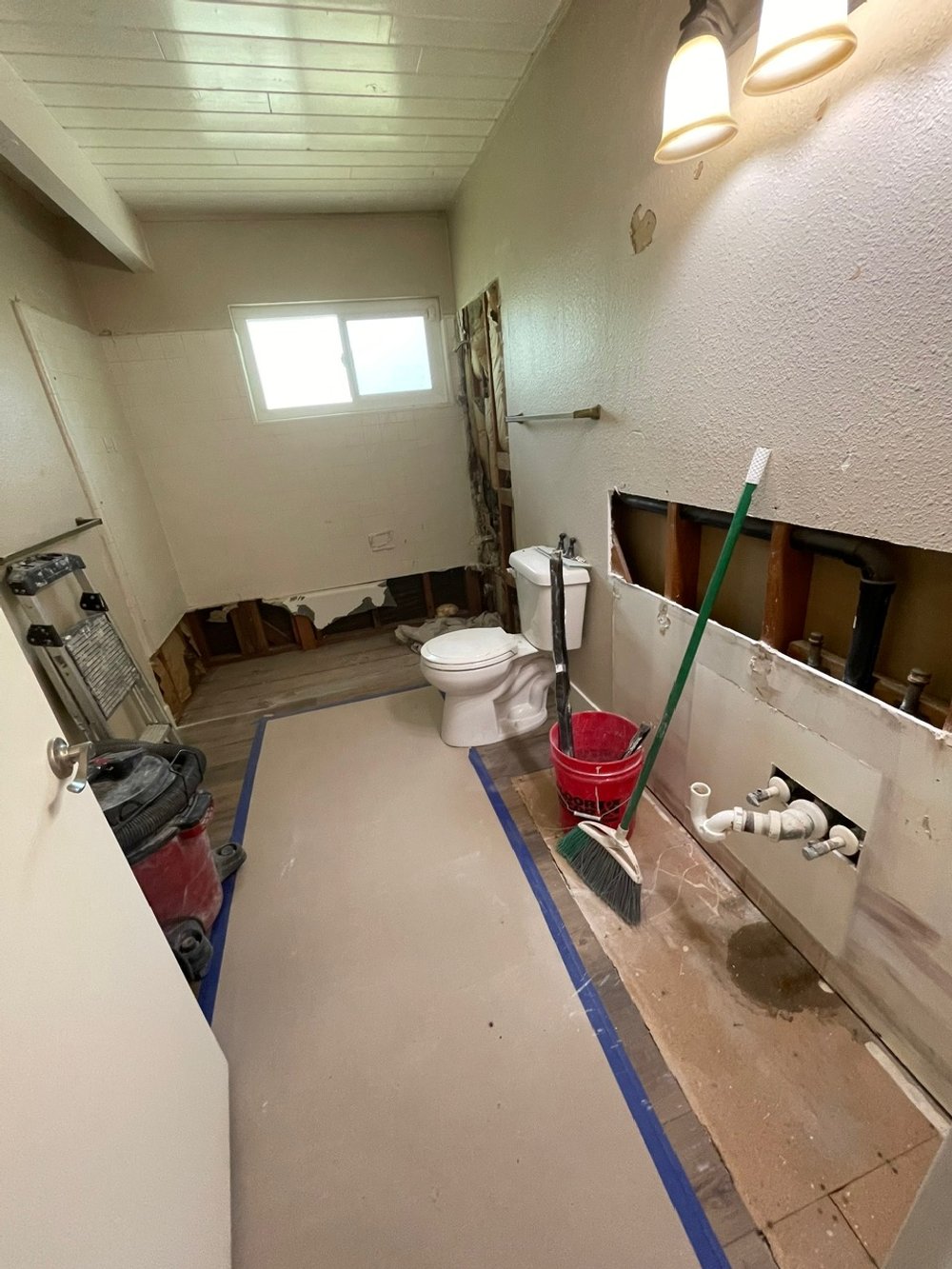 Bathroom under renovation with exposed wall pipes, a toilet, cleaning tools, and partially removed wall panels.