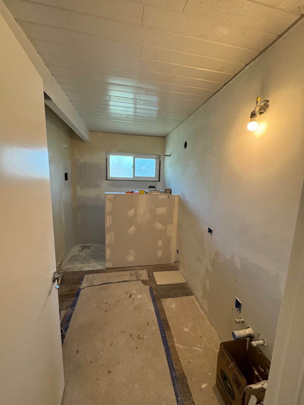 Small room under renovation with unfinished drywall, plywood, and construction tools, featuring a window and a single exposed light bulb on the wall.