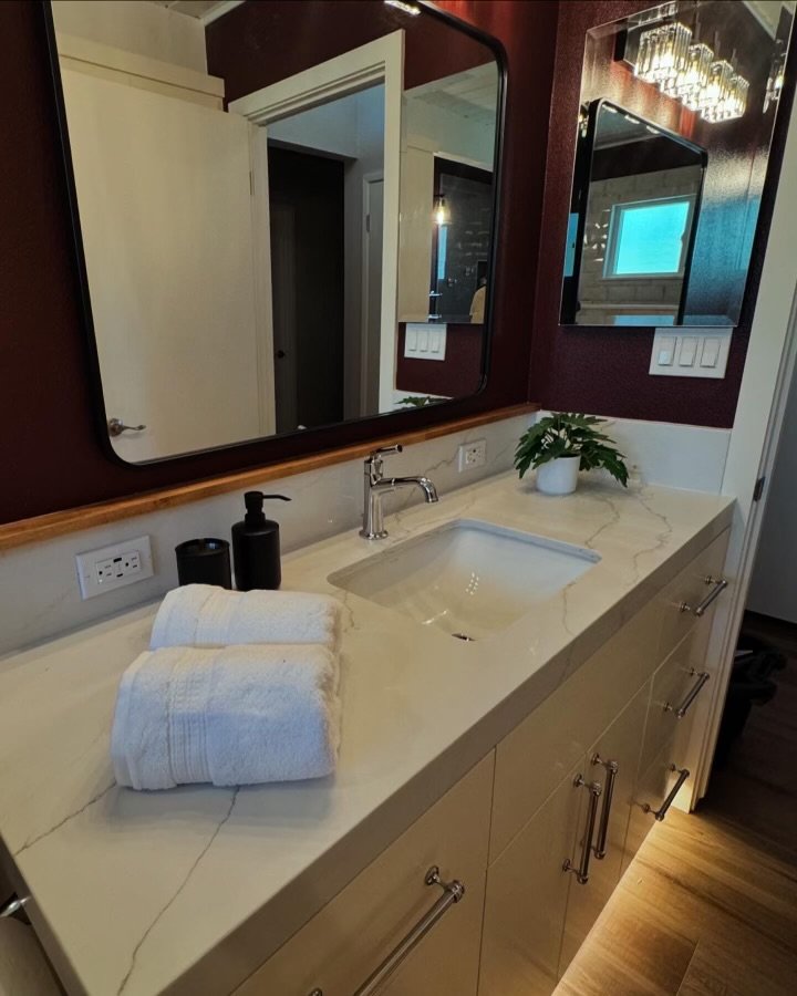 Bathroom vanity with a beige marble countertop, two rolled white towels, black soap and lotion dispensers, a plant, two large mirrors, and dark red walls.