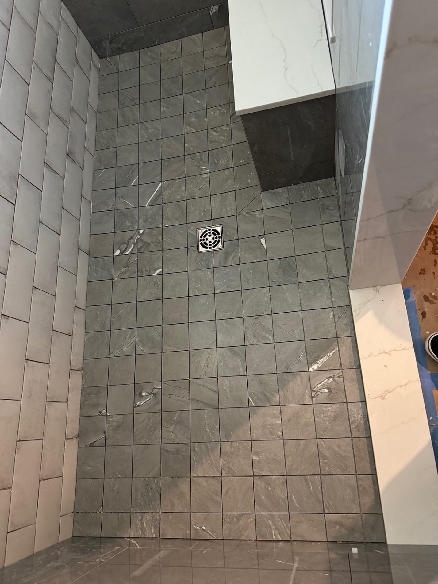 A tiled shower floor with grey marble tiles, a square drain in the center, and partially finished wall tiles on the sides.