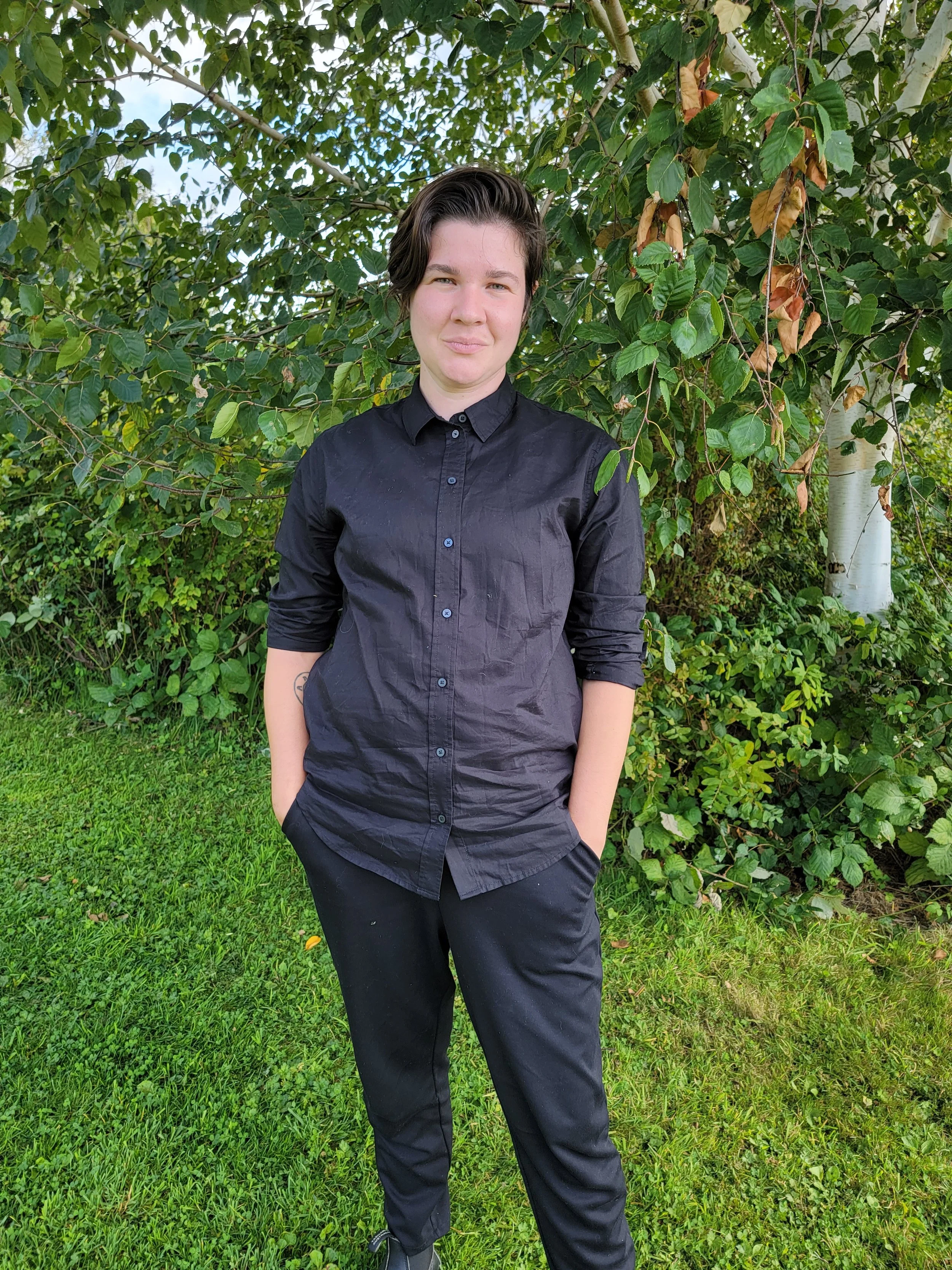 A non binary person with brown hair stands in front of a tree and bushes.