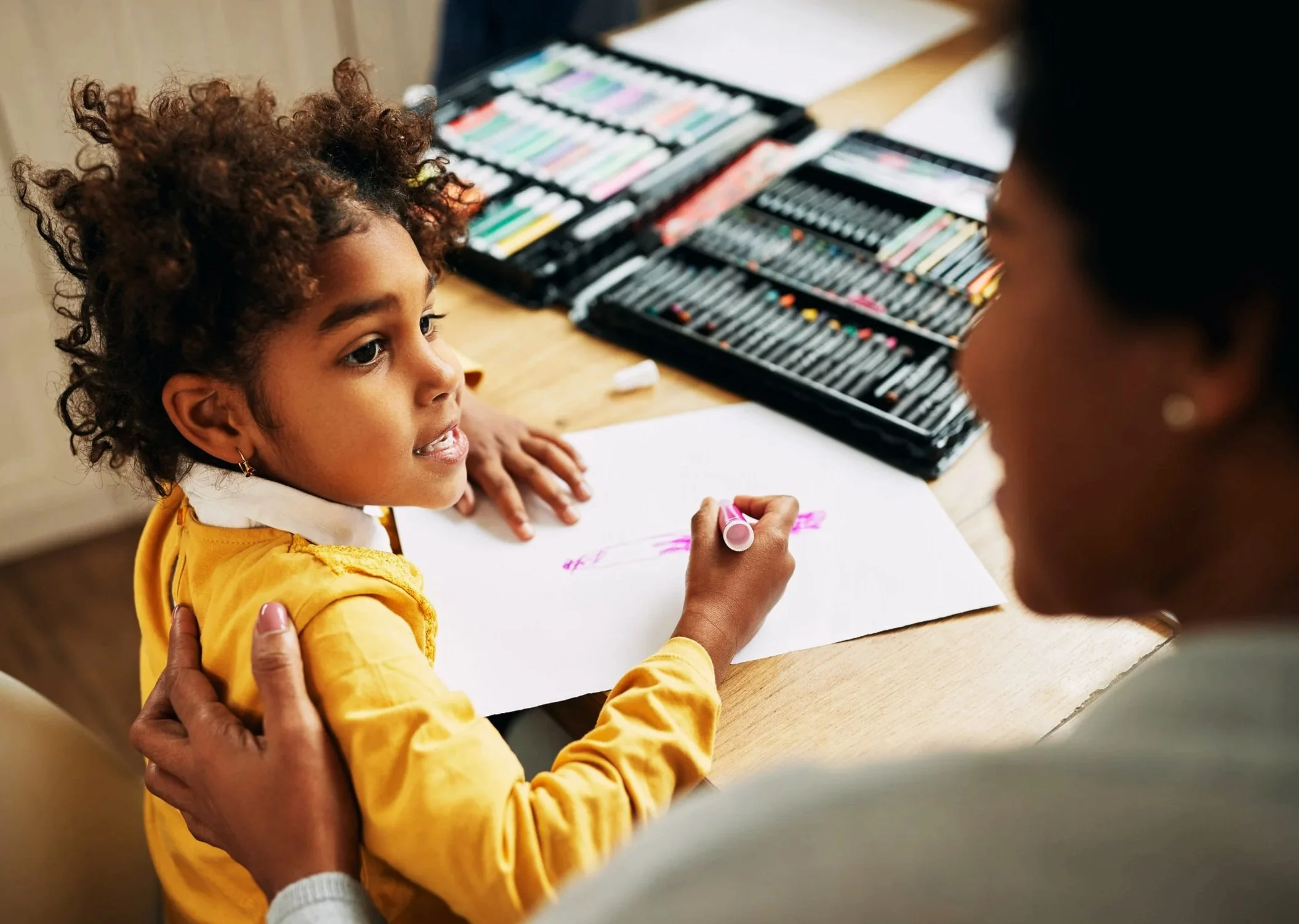 A young girl with curly hair drawing with a pink marker on a piece of paper, while an adult gently holds her shoulder. There are cases of colorful markers on the table.