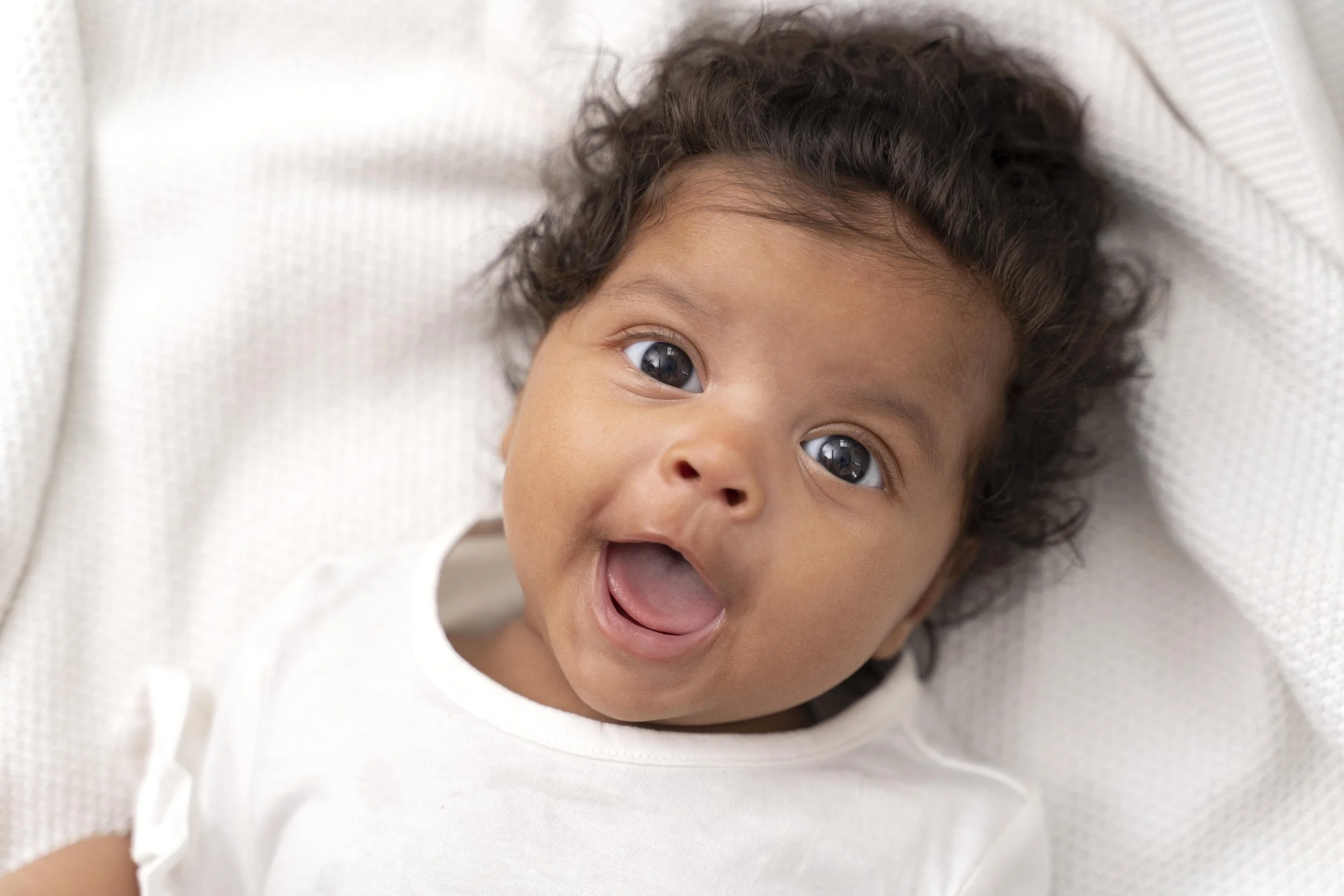 Close-up of a smiling baby lying on a white blanket, with dark curly hair and big, bright eyes.