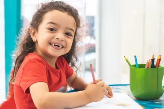 A young girl with curly hair smiling broadly, showing her teeth, against a teal background.