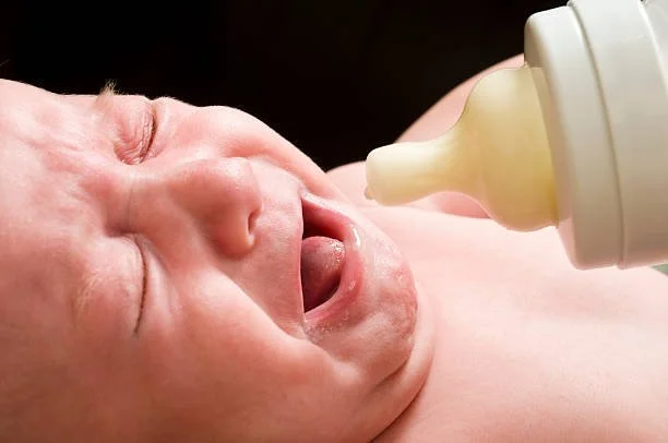 A black and white photograph of a baby being fed with a baby bottle, held by an adult's hand.