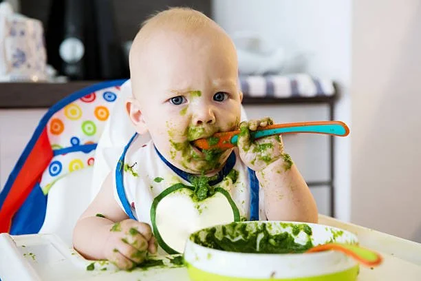 Baby with green food around mouth sitting in high chair, holding a spoon in hand, with a bowl of green food in front.
