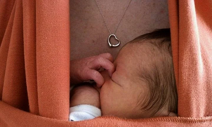 Close-up of a newborn baby breastfeeding, with a woman's chest and a heart-shaped necklace visible.