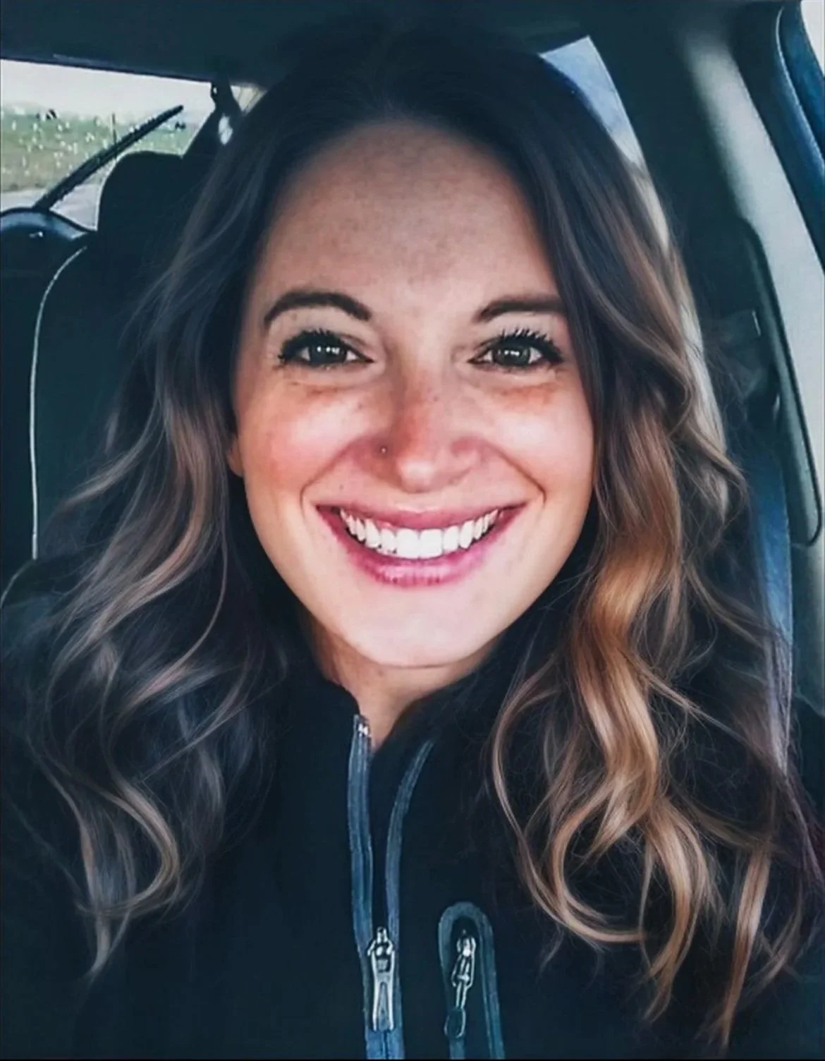 Close-up of a woman with long wavy brown hair smiling inside a vehicle.