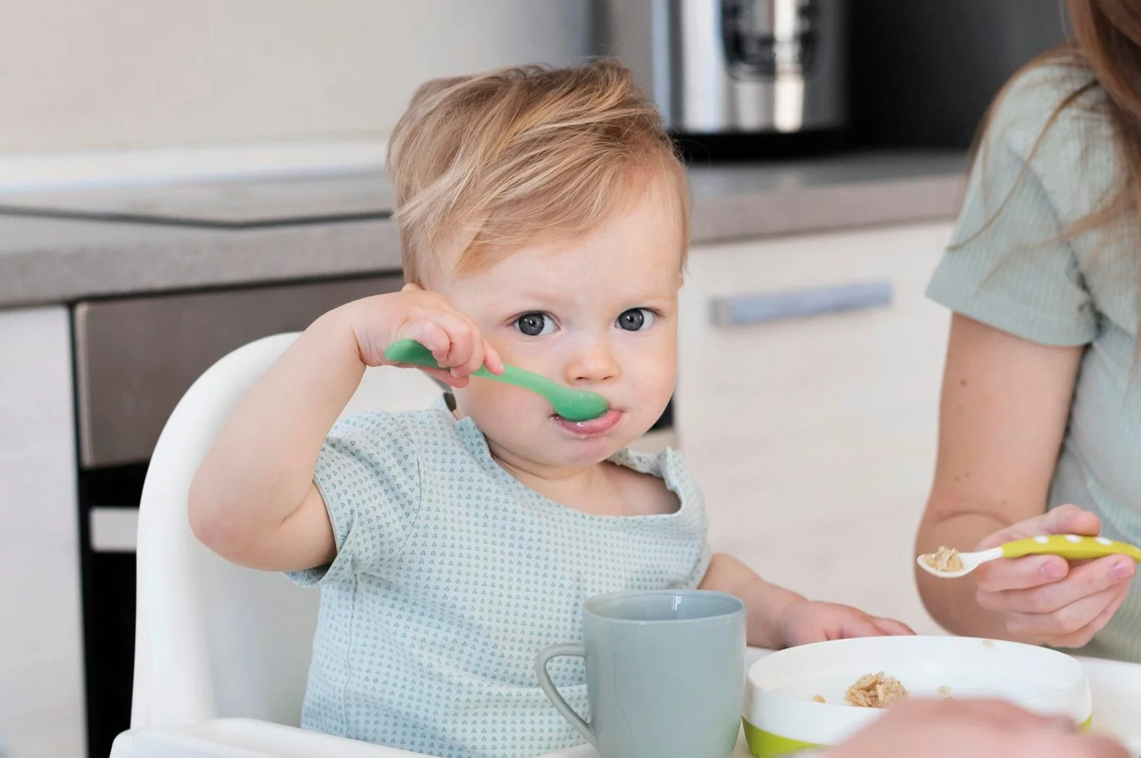 Young child with blonde hair and blue eyes sitting in a high chair, holding a green spoon near mouth with cereal and milk on plate and in a gray mug nearby, while a woman, partially visible, holds a spoon with cereal.