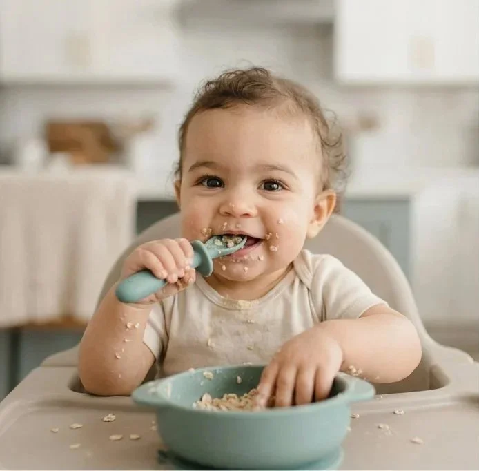 Black-and-white cartoon of a baby sitting cross-legged, holding a spoon near their mouth, with a bowl of food in front, smiling and winking.