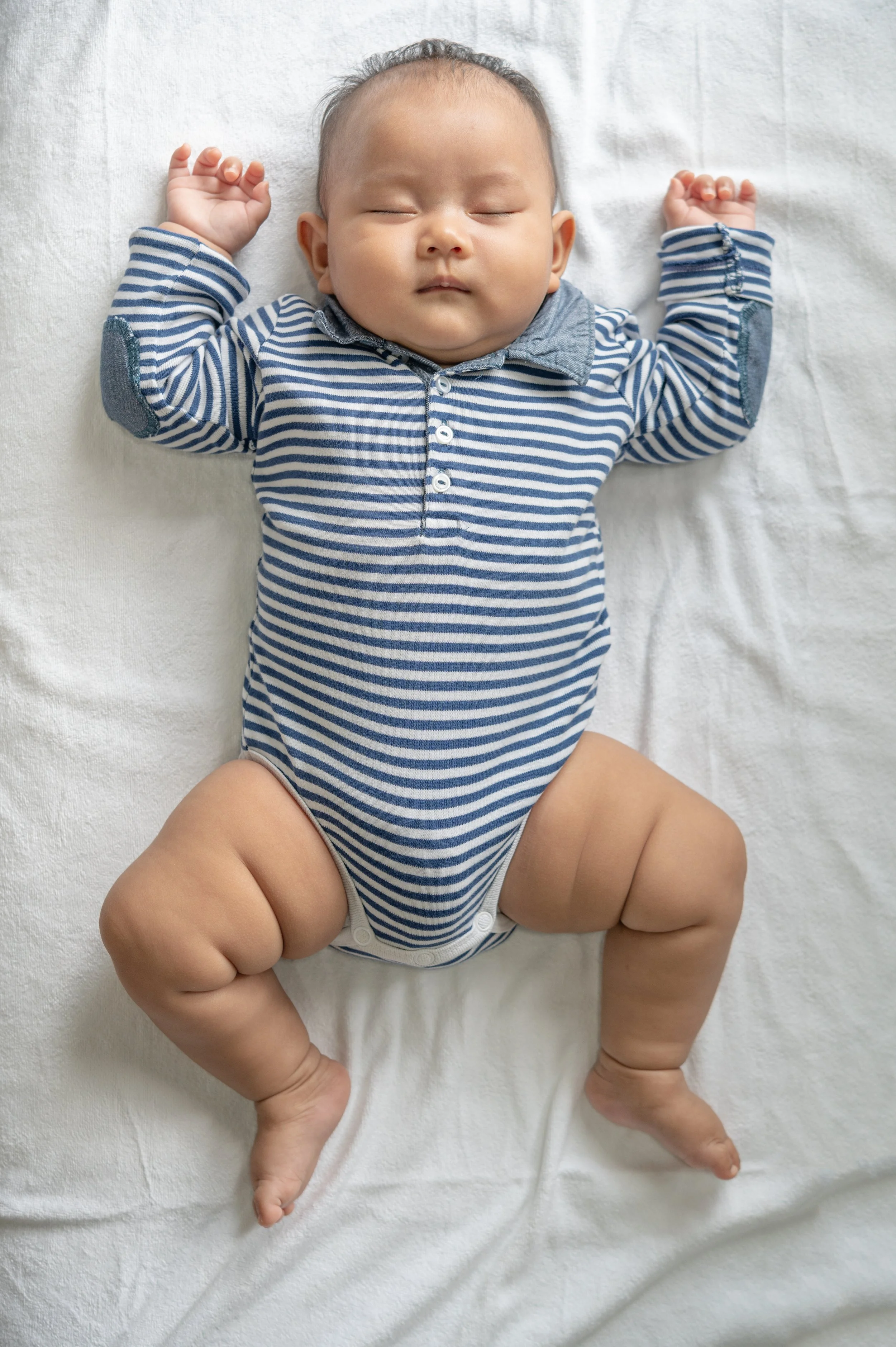 A sleeping baby lying on a white blanket, wearing a blue and white striped shirt and a diaper.