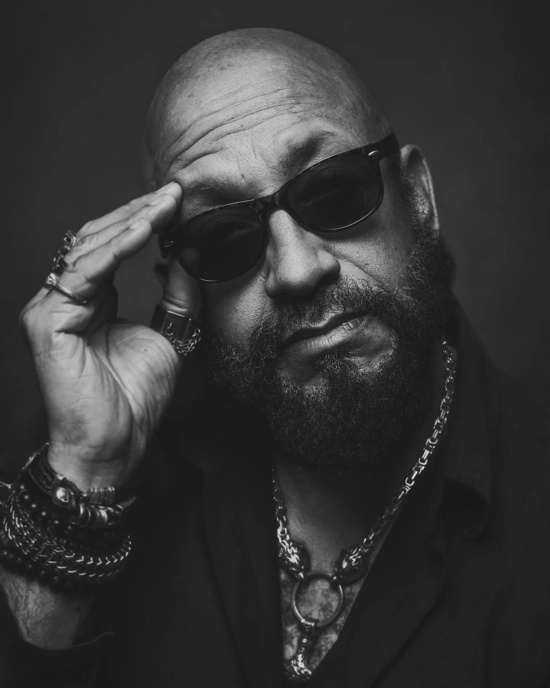Moody black and white portrait of a bald, black man with dark beard, wearing dark sunglasses.