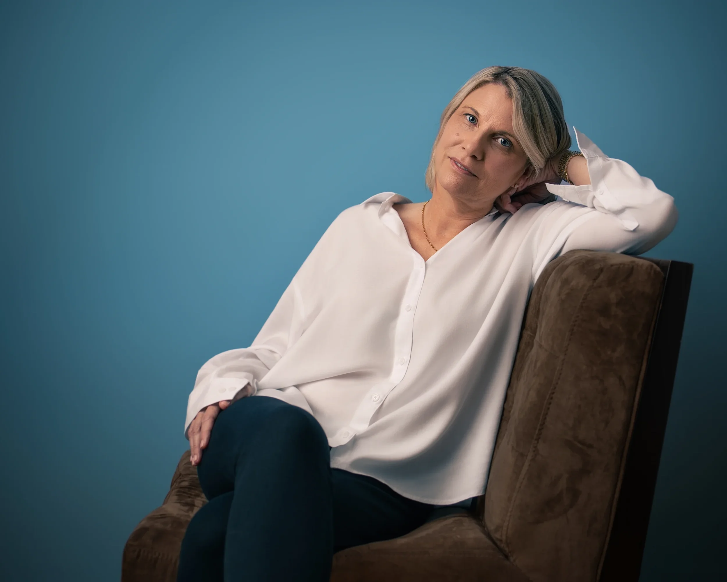A woman with blonde hair and blue eyes sitting on a brown sofa against a blue background. She is wearing a white button-up shirt and has a bracelet and a necklace. She is smiling softly and resting her head on her left hand.