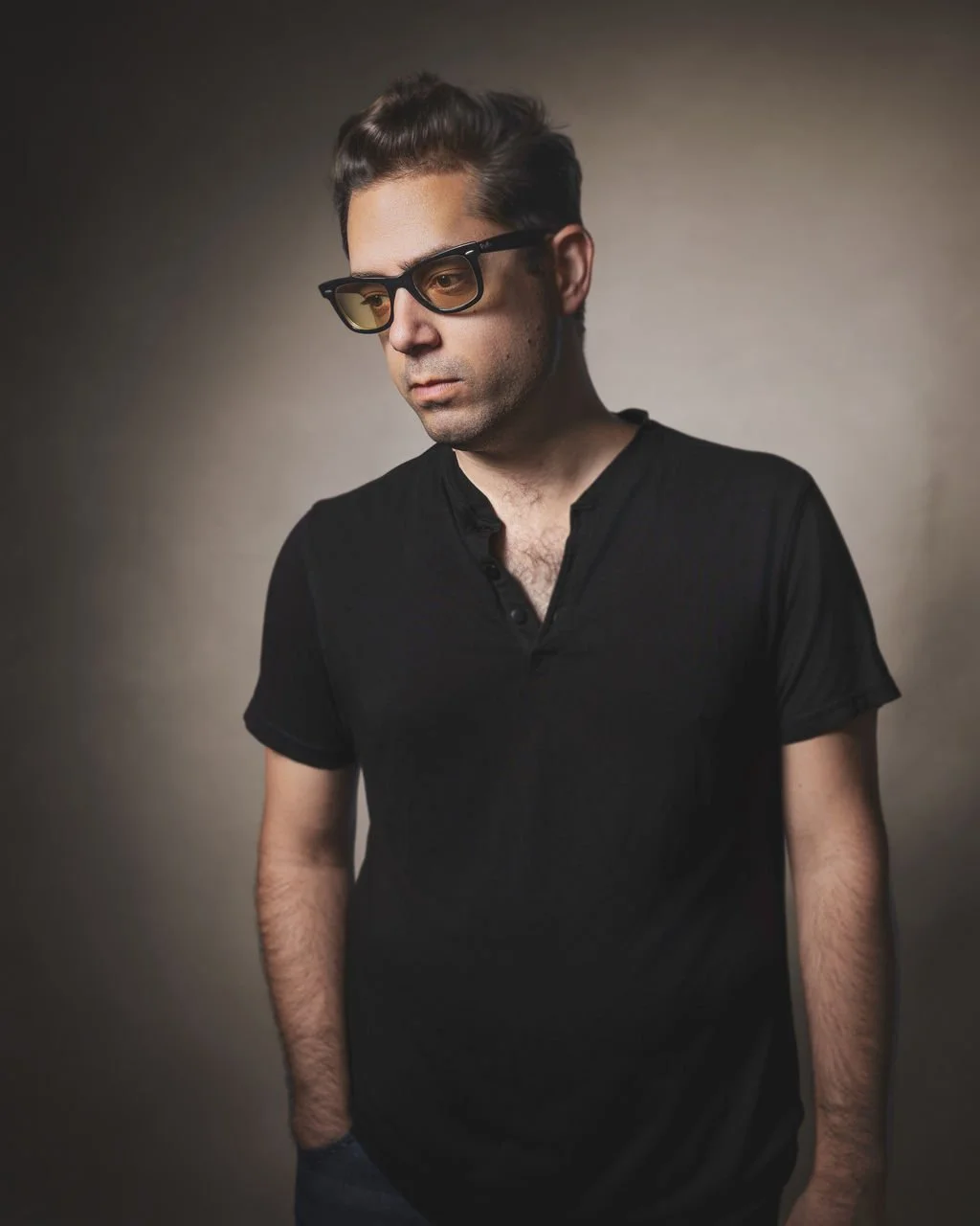 A young man with dark hair and glasses wearing a black shirt, looking down with a serious expression against a plain background.