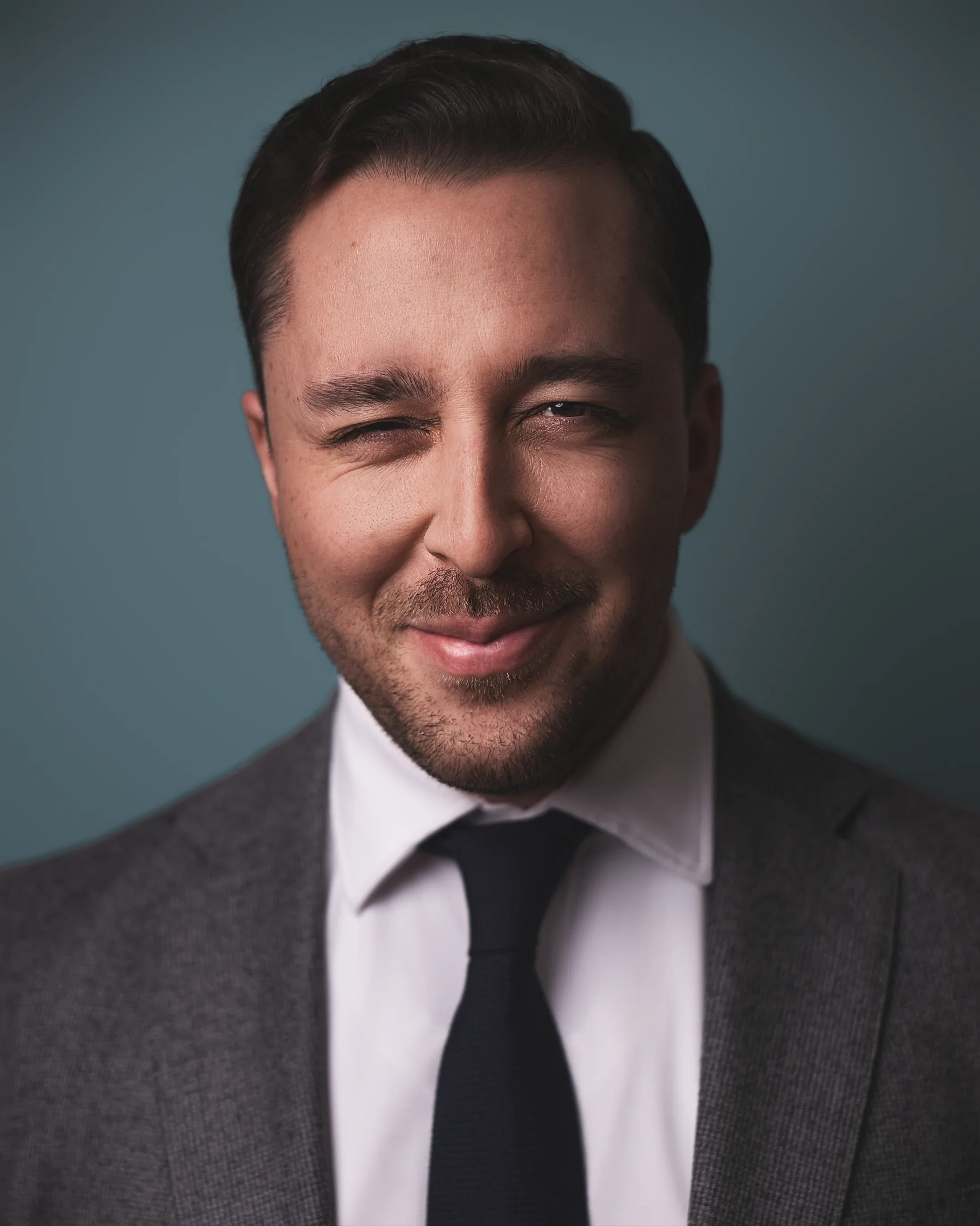 Portrait of a of a young man with beard stubble, wearing a grey suite and navy tie, smirking at the camera playfully.