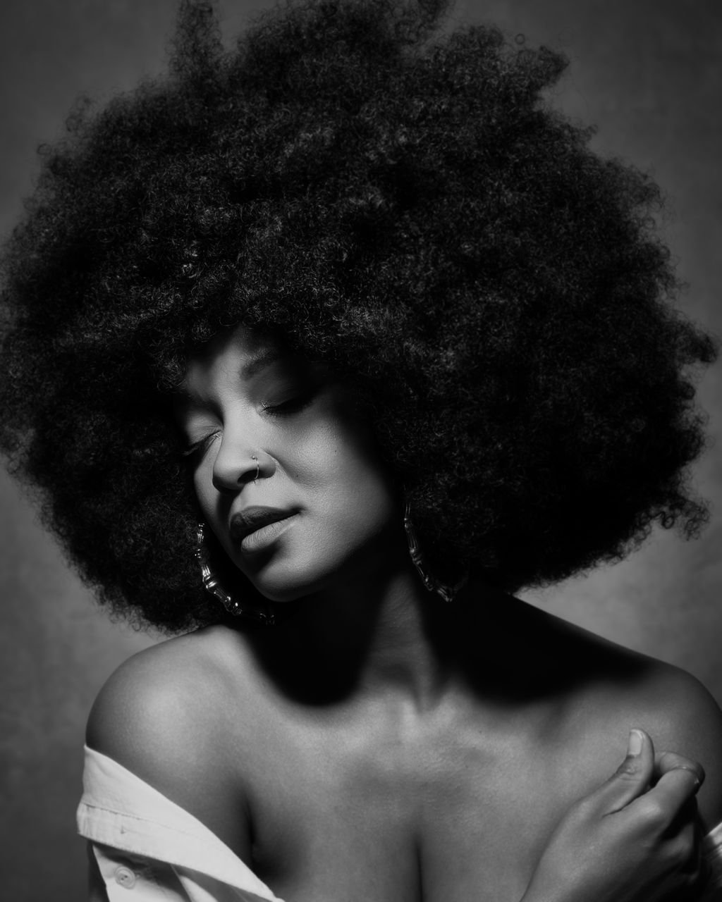 Black and white portrait of a woman with voluminous curly afro, her eyes closed, wearing earrings, with one hand resting on her shoulder, and a partially off-shoulder top.