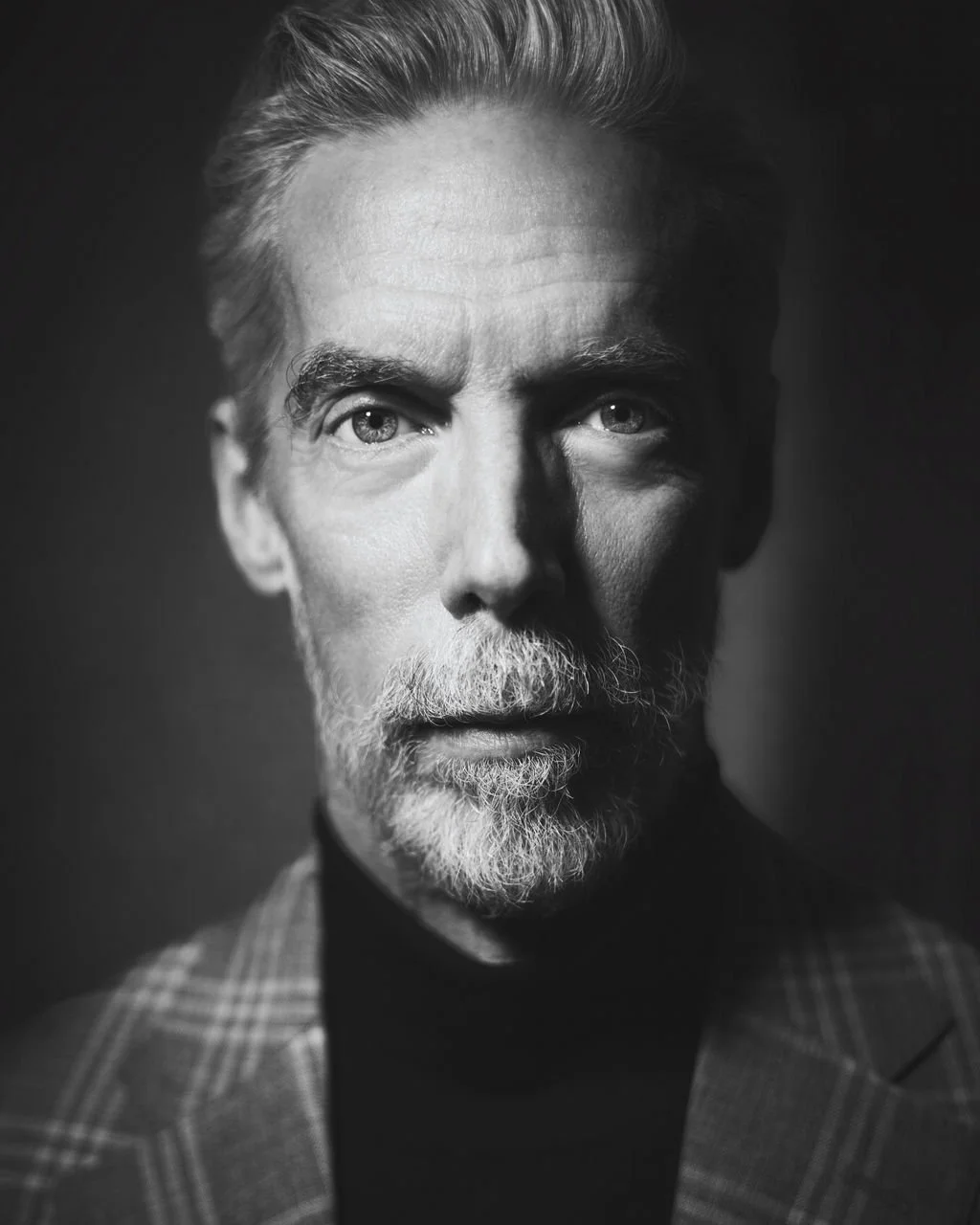 Close-up portrait of a mature man with gray hair, beard, and blue eyes, wearing a checkered blazer and black turtleneck, against a dark background.