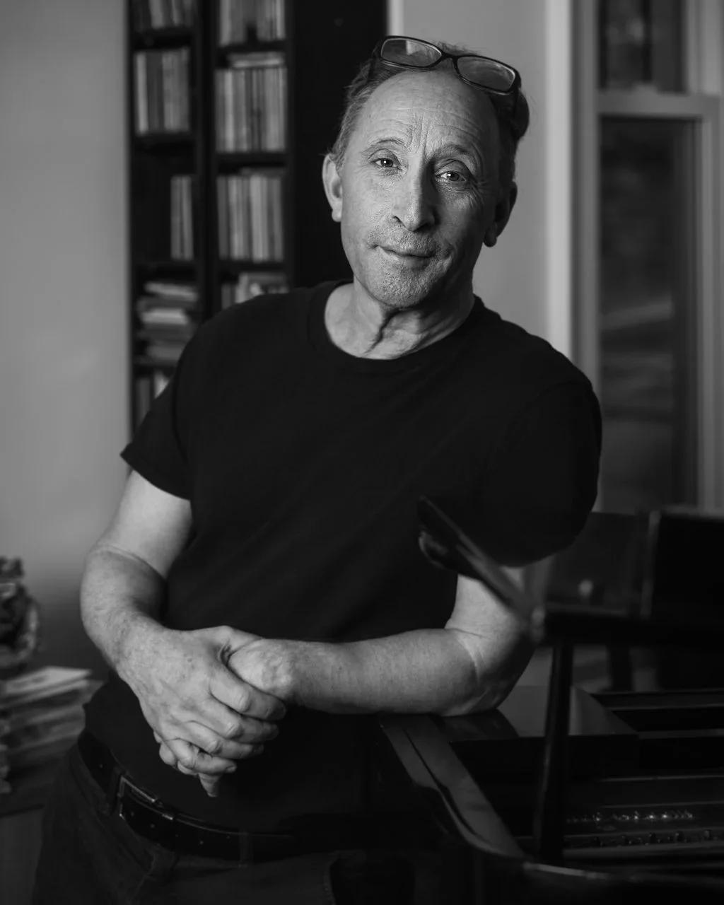 A black-and-white photo of a middle-aged man wearing a dark t-shirt and glasses perched on his head, standing near a piano with a blurred background of bookshelves.