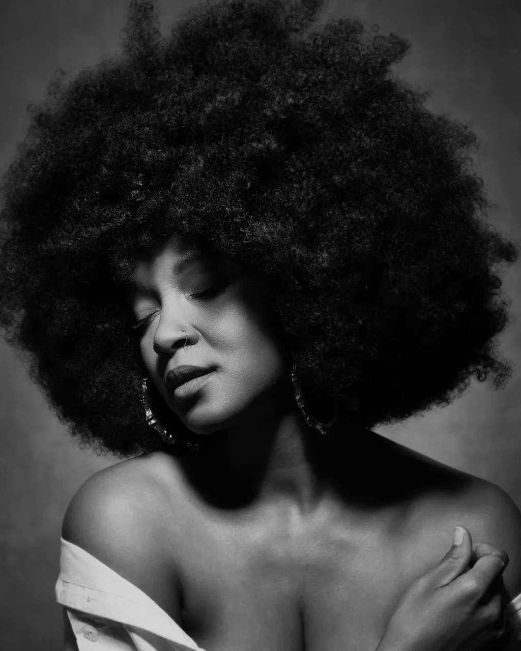 Black and white portrait of a woman with voluminous curly afro, her eyes closed, wearing earrings, with one hand resting on her shoulder, and a partially off-shoulder top.