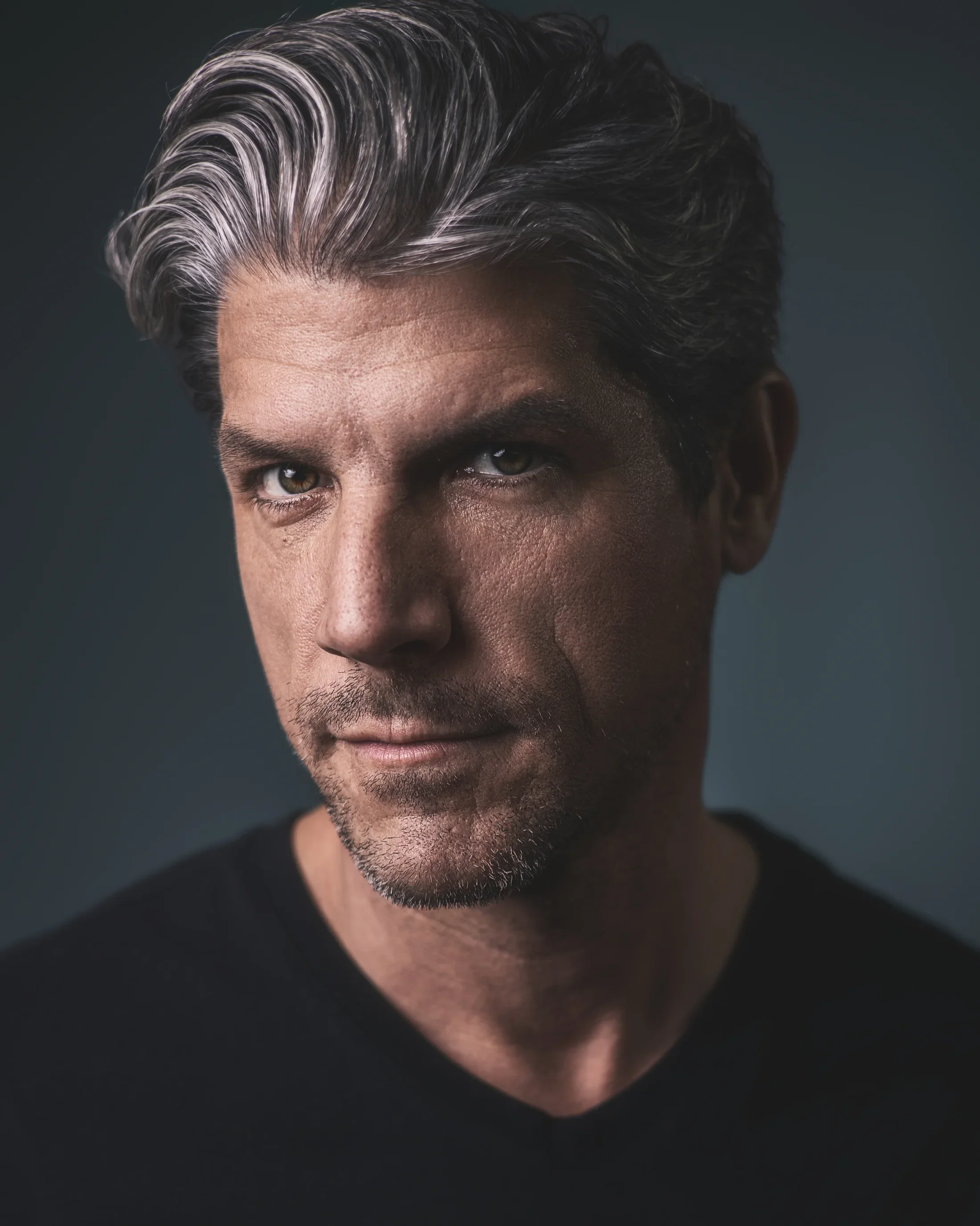 Close up portrait of a man with wavy salt and pepper hair wearing a black v-neck t-shirt.