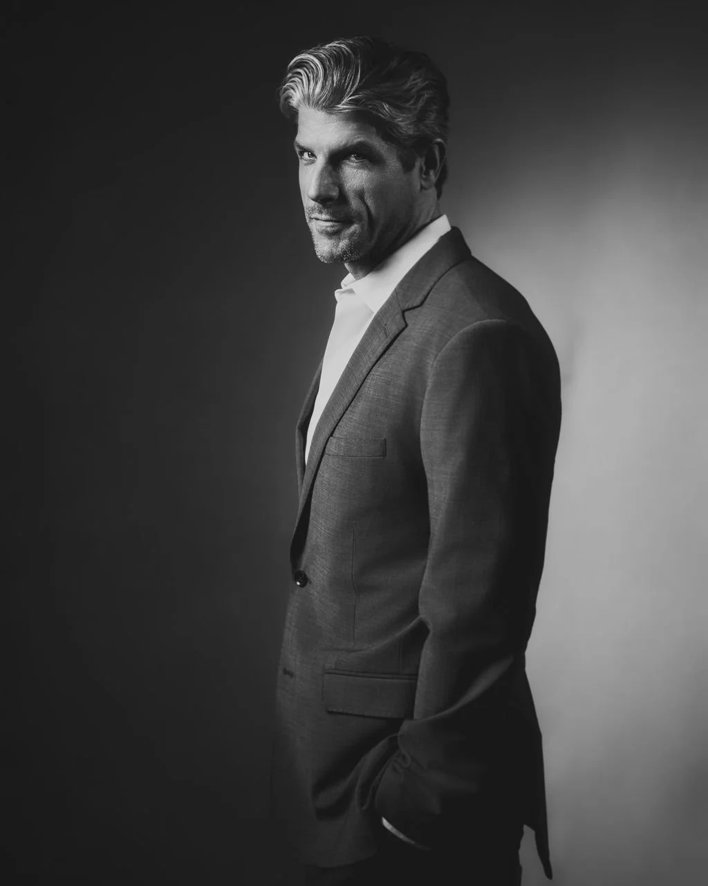 Black and white profile portrait of a man in a grey suit with a crisp white shirt and his hands in his pockets.