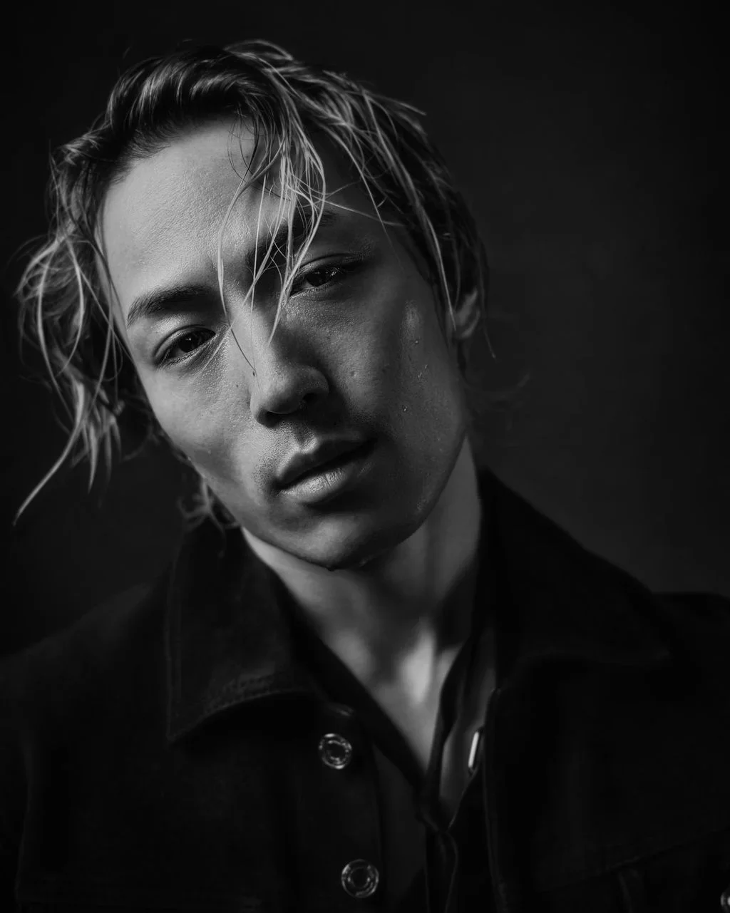 A black and white close-up portrait of a young Asian man with long, wet hair and a serious expression, wearing a dark jacket.