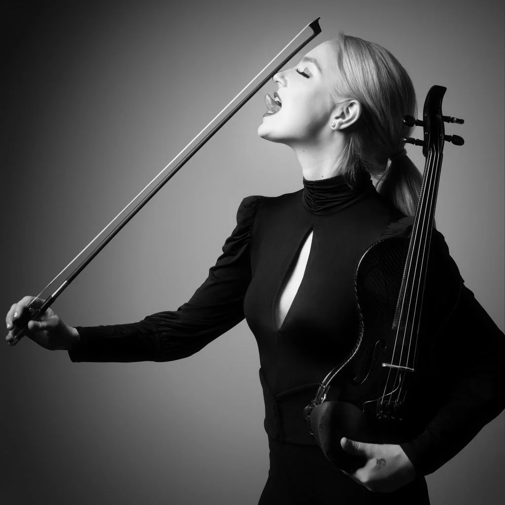 Black and white editorial portrait of violinist Emily Misura, playfully sticking her tongue out towards her bow.