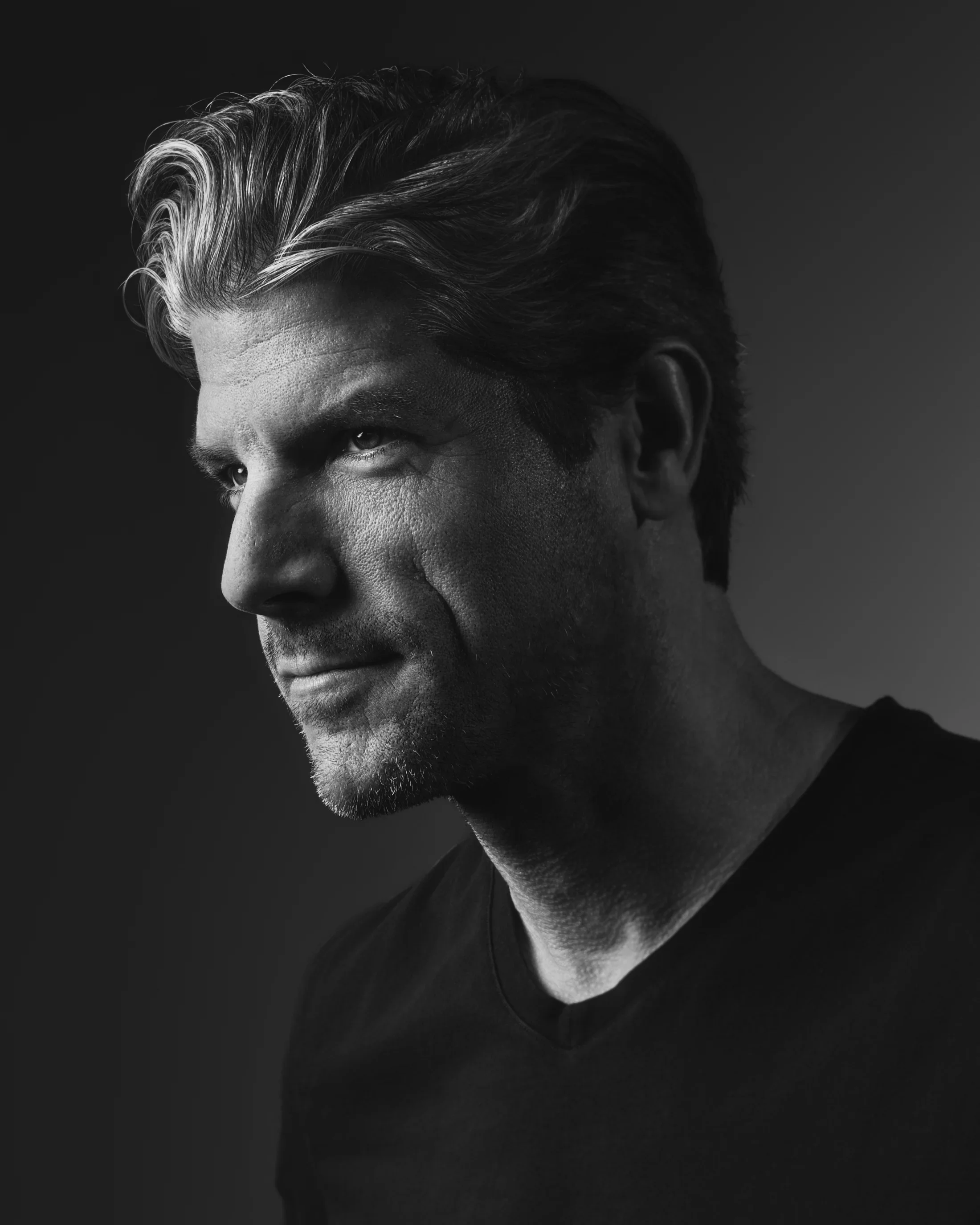 Black and white portrait of a man in a black t-shirt with wavy salt and pepper hair.