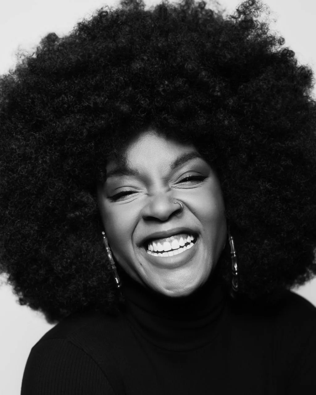 Black and white portrait of a black woman with voluminous curly afro, smiling broadly with her eyes closed, wearing a black turtleneck.
