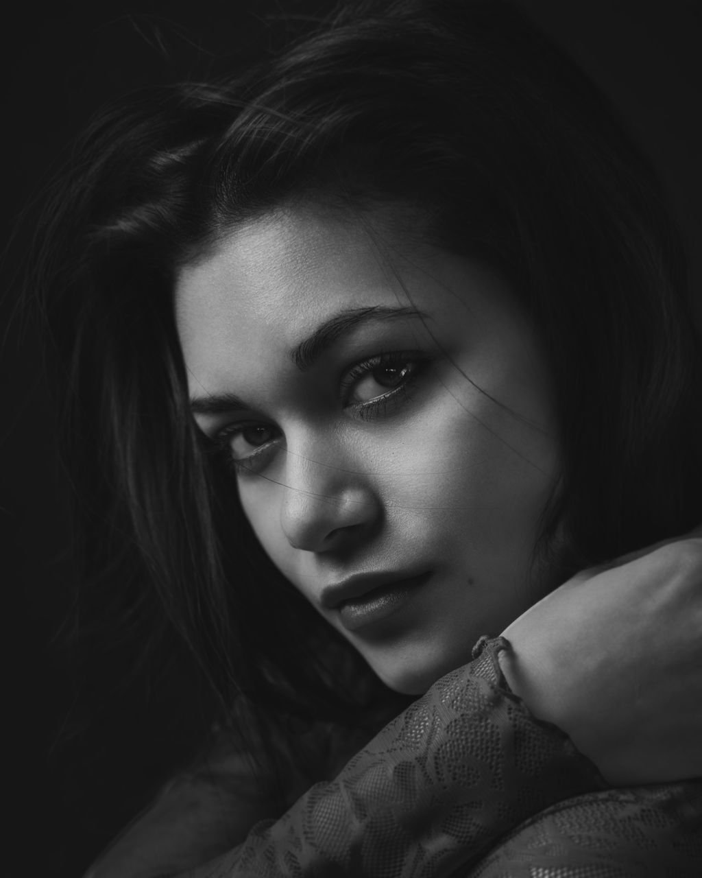 A black and white close-up portrait of a young woman with dark hair, intense eyes, and smooth skin, resting her chin on her hand.